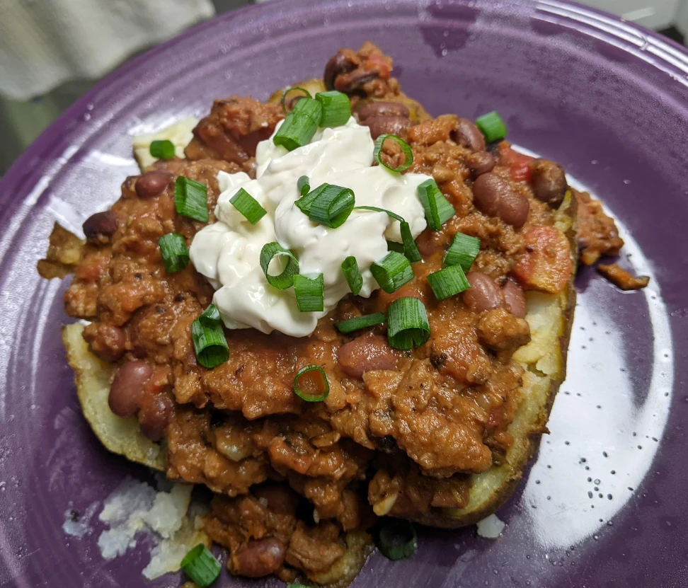 baked potato split in half and covered with a pile of chili, topped with a dollop of what looks like sour cream and garnished with green onions on a purple Fiestaware plate