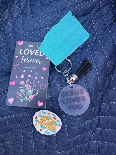 A card with the text you are loved forever and Romans 8:38 a blue prayer cloth, a small keychain with the text “Jesus loves you” and a sticker which reads “wake up and choose positivity” 