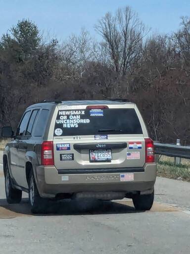 A tan Jeep Patriot with letters that spell out "NEWSMAX OR OAN UNCENSORED NEWS" A thin blue line sticker that is illegible, a "Thank God" Sticker, a Trump sticker, and some other things I can't make out. Would it be too malicious to add the license plate number here as well? 